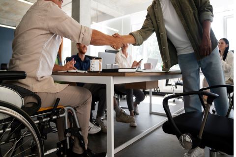 Handschlag auf der Arbeit, ein Mann steht, der andere sitzt im Rollstuhl, Kollegen applaudieren