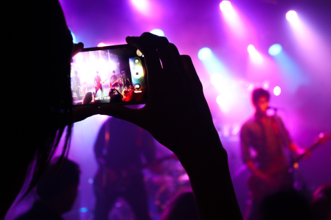 Auf dem Bild ist zu sehen, wie eine Person auf einem Konzert ist und mit dem Smartphone ein Foto von den Künstler:innen auf der Bühne macht
