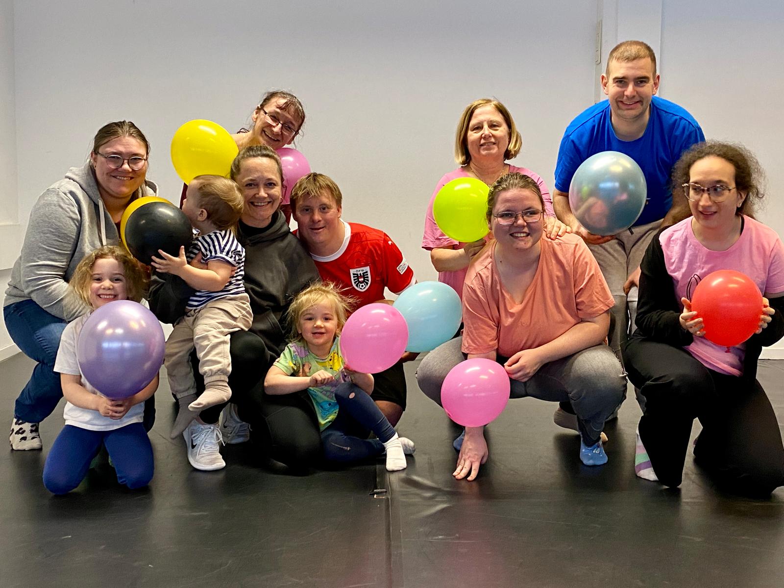 Das Bild zeigt zeigt lächelnde Personen in unterschiedlichem Alter, auch Kinder sind dabei. Alle haben einen Luftballon in der Hand und sitzen, knien oder hocken am Boden.