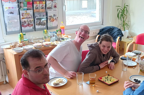 Das Foto zeigt Geburtstagskind Alexander, wie er der Kamera die Zunge herausstreckt. Er sitzt mit zwei weiteren TBW Kunden um ein Mensch Ärger Dich Nicht Spiel.