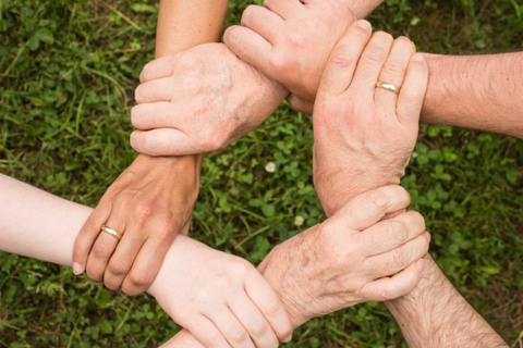 Auf dem Beitragsbild sieht man verschiedene Hände, die sich im Kreis jeweils am Handgelenk fassen und somit ein starkes Team bilden.