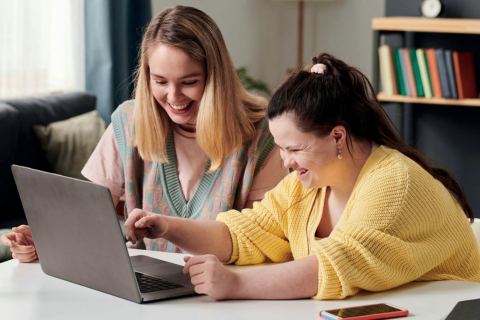 Eine junge Frau ohne Behinderung und eine junge Frau mit Behinderung sitzen gemeinsam vor einem Laptop und schauen fröhlich lachend auf den Bildschirm. Die junge Frau mit Behinderung tippt auf der Tastatur.