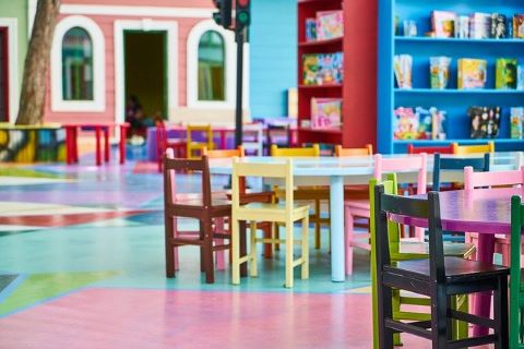 Gruppenraum in einem Kindergarten, in dem Tische mit Sesseln zu sehen sind, Regale mit unterschiedlichen Spielmaterialien.