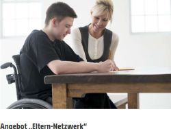 Ein junger Mann im Rollstuhl sitzt an einem Tisch und liest einen Zettel. Ein Dame sitzt neben ihm und bespricht mit ihm, was aus dem Zettel steht. 