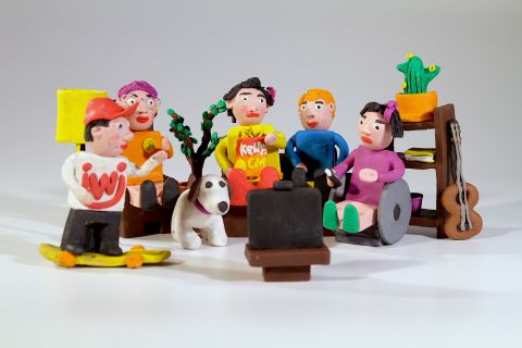 Das Bild zeigt eine Gruppe aus Knetfiguren, es ist ein Screenshot aus dem iwi-Imagefilm. Das Maskottchen Imi steht auf seinem Skateboard mit einer Gruppe anderer vor einem Fernseher.