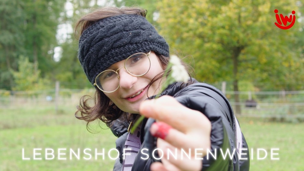 Eine Frau lächelt und hält eine Blume in die Kamera. Im Hintergrund sind Bäume und viel Grün.