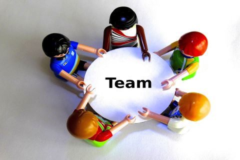 Blick von oben auf fünf bunte unterschiedliche Playmobilfiguren. Diese sitzen um einen weißen, runden Tisch auf dem das Wort Team in schwarzen großen Buchstaben geschrieben steht.